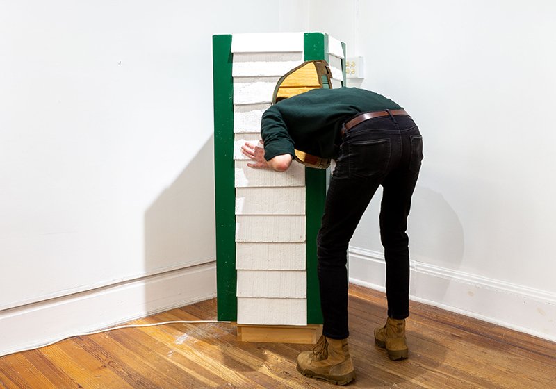A person bent down and sticking there head into a hole in a 4' section of a white and green chimney swift tower